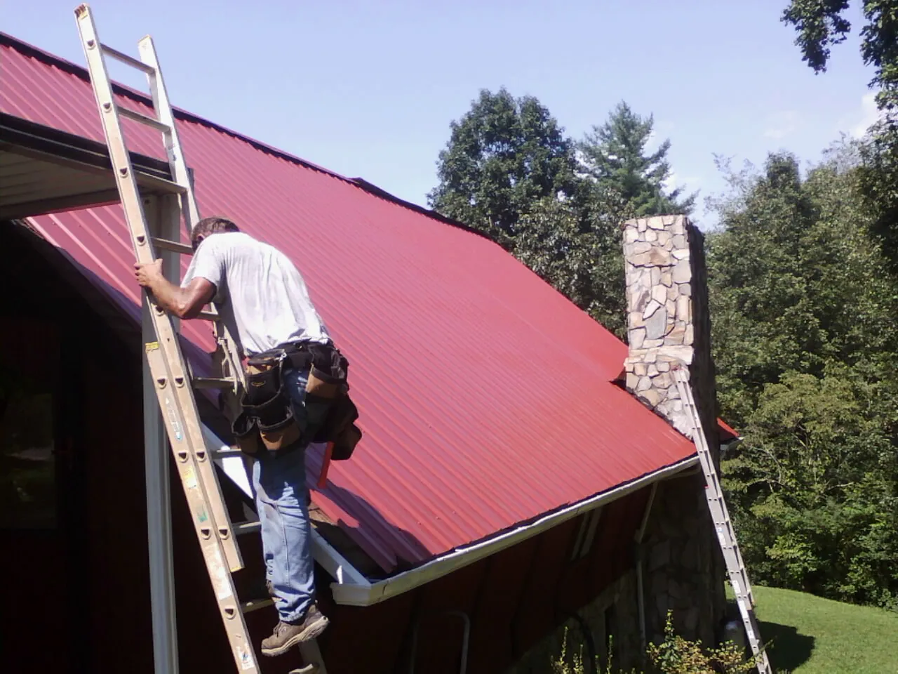 Roofer on a ladder beside a red metal roof and stone chimney — Soffit & Fascia Repair by Roofing Brothers in Kernersville